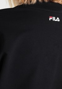 Vista trasera de una persona que lleva una sudadera negra de FILA con cuello de canalé y el logo blanco-rojo de FILA cerca del escote.