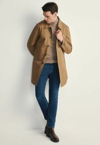 Beżowy płaszcz wodoodporny z kołnierzem, zapięciem na guziki i bocznymi kieszeniami, noszony na beżowy golf i niebieskie dżinsy, zestawiony z brązowymi botkami do kostek.