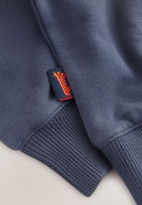 Mangas de sudadera azul marino con puños acanalados y una pequeña etiqueta azul marino con texto bordado en rojo.