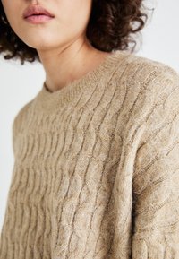 Beige kabelstrikket genser med ribbestrikket hals og teksturert mønster, laget av mykt, lodden materiale. Nærbilde som viser detaljer i stoffet.