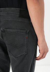 Black denim jeans with a smooth texture, featuring a back pocket and a subtle red logo accent. The waistband has a black leather patch.