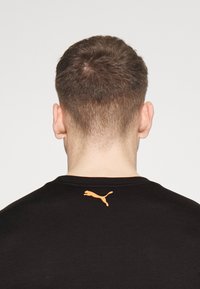 Camiseta negra con cuello redondo, que presenta un logo naranja de Puma en la parte superior de la espalda. El tejido parece suave y ligero.