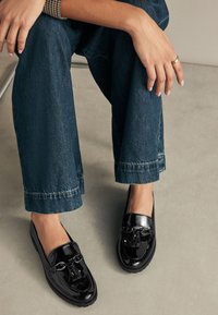 Mocassins en cuir verni noir à finition lisse, ornés de glands décoratifs et d'une semelle épaisse, associés à un jean large bleu foncé.