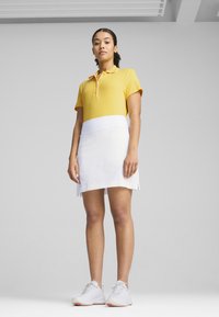 Polo giallo con colletto e maniche corte, abbinato a una gonna bianca. Presenta un design aderente e scarpe da ginnastica grigie.