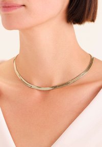 Collana argentata con un design piatto e flessibile, caratterizzata da motivi a zigzag. Texture liscia con una leggera lucentezza, posata sulla pelle nuda.