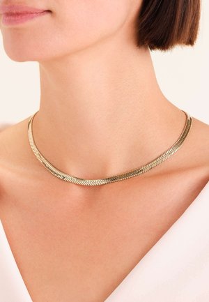 Collana argentata con un design piatto e flessibile, caratterizzata da motivi a zigzag. Texture liscia con una leggera lucentezza, posata sulla pelle nuda.