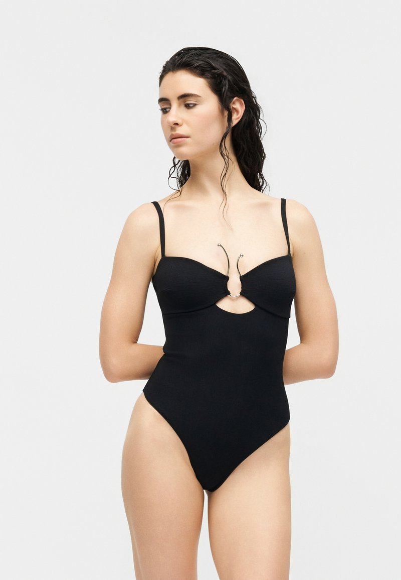 Femme aux cheveux noirs mouillés porte un maillot de bain une pièce noir avec de fines bretelles et une découpe avec des détails métalliques au niveau du buste.