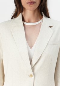 Kremfarget lin blazer med én knapp, notched revers og brystlomme, lagt over en lys mesh-top. Teksturlignende stoff synlig.