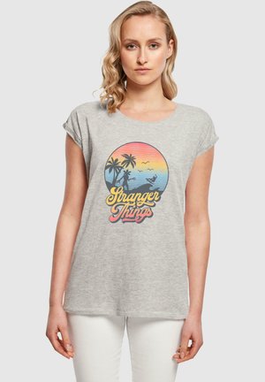 Grå t-skjorte med kort erm som har et sirkulært grafisk motiv med palmer, en solnedgangsgradients og "Stranger Things"-tekst i en fet skrift.