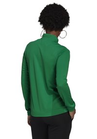 Grön långärmad topp med hög krage och jämn textur. Har en figurnära design och når till midjan, i kombination med svarta byxor.