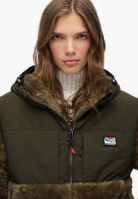 Padded jacket in olive green with a faux fur collar and sleeves, featuring a zipper and red accent pull, branded patch on the chest.