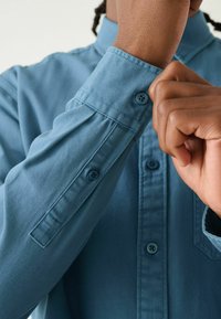 Lichtblauw, overhemd met lange mouwen en een knoop-sluit ontwerp. Heeft een gestructureerde kraag en knoopaccenten op de manchetten. Katoenen textuur zichtbaar.