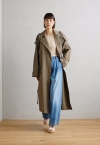 Gabardina caqui con mangas anchas, superpuesta sobre una blusa beige, combinada con unos pantalones vaqueros de pierna ancha azul claro y sandalias de plataforma blancas. Texturas suaves.