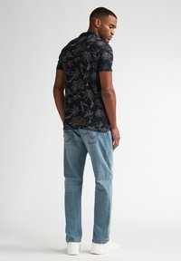 Marineblå polo med gyldent blomsterprint, korte ærmer, kombineret med lyseblå denimjeans og hvide sneakers. Set bagfra.