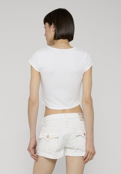 Femme aux cheveux courts et foncés, debout de dos, portant un t-shirt court blanc et un short en jean blanc avec des poches à l'arrière.