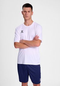 Valge lühikeste varrukatega sportlik T-särk, mille rinnal on must logo. Sobitatud siniste lühikestega sportliku välimuse loomiseks.