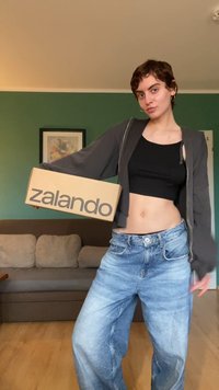 Caixa de cartão com "zalando" impresso em cinza, segurada por uma pessoa que veste um top preto cropped e um hoodie cinza de fecho, combinado com calças largas azuis claras.