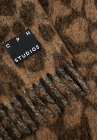 Écharpe léopard brun et beige avec un bord frangé, en tissu doux, portant une étiquette noire avec du texte blanc : "CPH STUDIOS."
