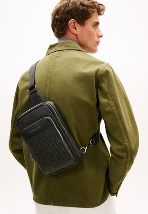 Schultertasche aus schwarzem, strukturiertem Material mit geometrischen Mustern, verstellbarem Riemen und Reißverschlussdetails, quer über einer olivgrünen Jacke getragen.