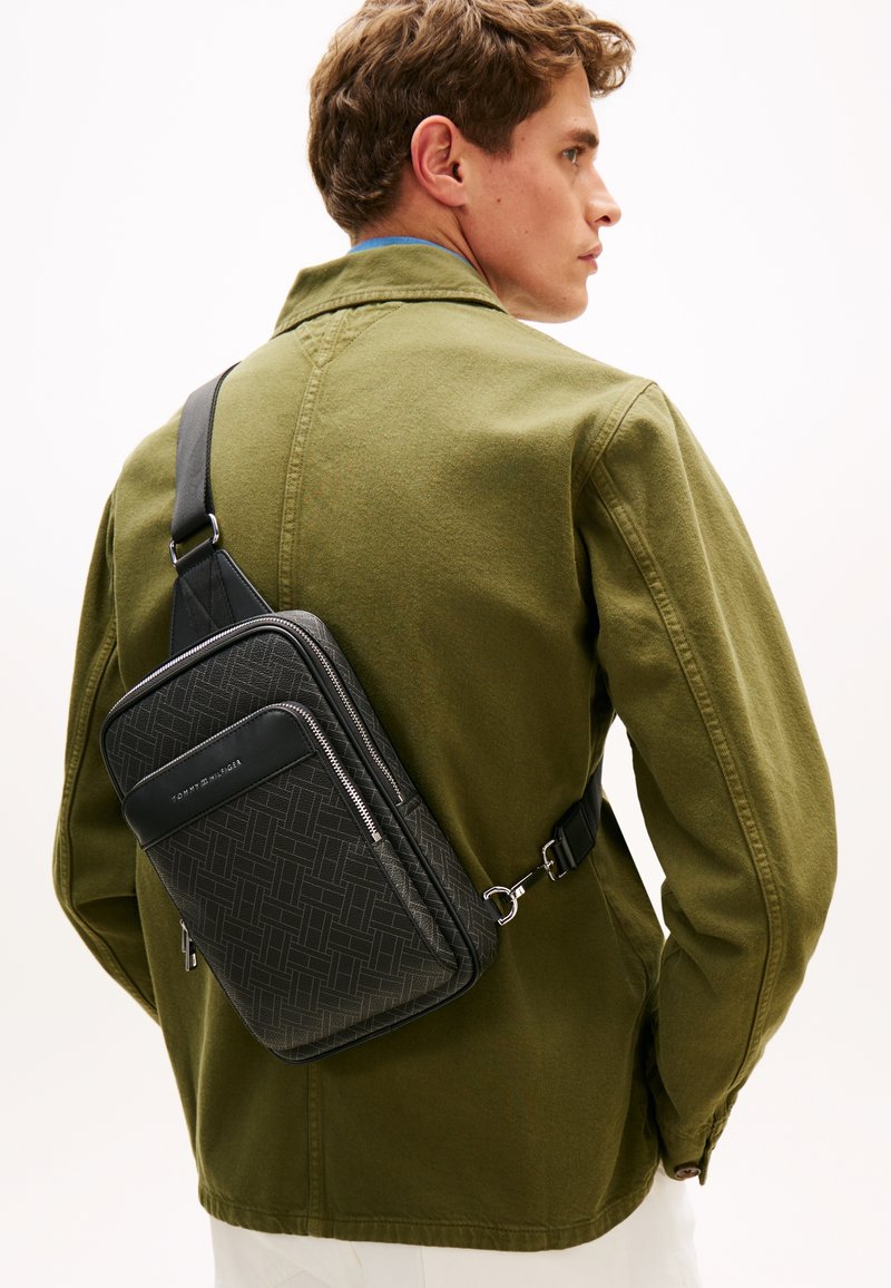 Τσάντα ώμου από μαύρο υλικό με υφή, με γεωμετρικά μοτίβα, ρυθμιζόμενο λουράκι και λεπτομέρειες με φερμουάρ, φορεμένη χιαστί πάνω από ένα ελαιώδες μπουφάν.