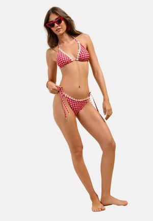 Jonge vrouw draagt een rood-wit geruit bikini met madeliefjes afwerking, rode cat-eye zonnebril, en houdt de zijstrik van de bikini vast tegen een effen achtergrond.