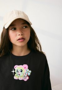 Czarny t-shirt z kolorową grafiką w formie rysunkowego misia, który ma kwiat i wisienki. Czapka jest beżowa, wykonana z materiału, z zaokrąglonym daszkiem.