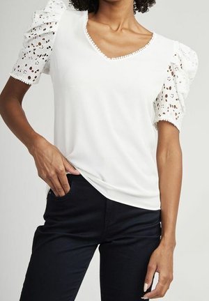 Kobieta w białej bluzce z dekoltem w kształcie litery V, z koronkowymi bufkami na rękawach i czarnych spodniach, z ręką wsuniętą w prawą kieszeń, na jednolitym tle.