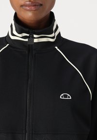 Crna jakna s patentnim zatvaračem i visokim ovratom, koja ima bijele prugaste detalje, teksturiranu površinu i mali izvezeni logo na prsima.