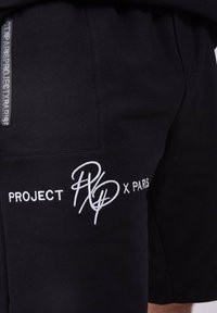 Pantalones cortos negros hechos de una tela suave con el texto bordado en blanco "PROJECT P x PARIS" y una etiqueta decorativa negra en el costado.