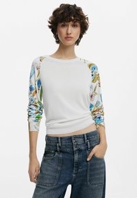 Pull en tricot blanc avec des manches à motifs présentant des designs floraux colorés. Le corps est lisse, tandis que les manches sont texturées avec des couleurs mixtes.