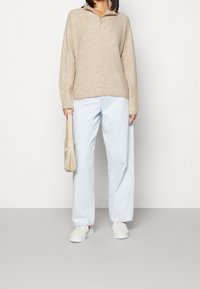 Person, die einen beigen Strickpullover, hellblaue weit geschnittene Jeans und weiße Sneaker trägt und eine beigen Handtasche vor einem schlichten Hintergrund hält.
