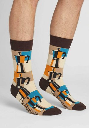 Nogi w beżowych skarpetach z ciemnobrązowymi ściągaczami i palcami, zdobione kolorowymi geometrycznymi wzorami kawiarek moka w kolorach niebieskim, pomarańczowym, czarnym i beżowym.