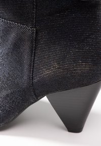 Botte de cheville texturée noire avec une finition métallique. Présente un talon en forme de cône et un design épuré, mettant en valeur son tissu brillant.