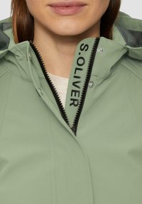 Chaqueta impermeable de color verde claro con cremallera frontal y texto negro, además de broches. El material es suave con un acabado mate.