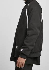 Chaqueta negra con cremallera, detalles blancos en los laterales, puños acanalados y dobladillo redondeado. Presenta un parche con logo en el lateral inferior y una textura suave.