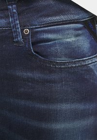 Jeans de mezclilla azul oscuro con un bolsillo frontal, remache plateado, patrones de desvanecimiento sutiles y costuras visibles a lo largo de los bordes.
