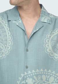 Helles blaues Kurzarmhemd mit Kragen, das ein großes weißes Paisleymuster und eine vordere Knopfleiste aufweist.