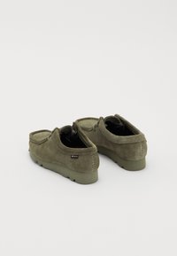 Chaussures en daim vertes avec un design bas et arrondi, des détails cousus, une semelle en caoutchouc et des étiquettes sur le côté. La vue arrière montre un contraste de couleur interne.