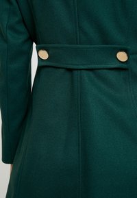 Cappotto verde in lana caratterizzato da un design strutturato, con un dettaglio cintura fissato da due bottoni dorati sulla schiena. Texture liscia e cuciture sartoriali.