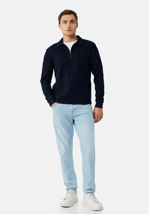 Mężczyzna w granatowym swetrze z krótkim zamkiem, białej koszulce, jasnoniebieskich jeansach i białych trampkach, stojący z jedną ręką w kieszeni.