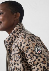 Chaqueta acolchada en beige con estampado de leopardo en negro y marr ón. Incluye cierre de cremallera y un parche redondo con logo en la manga izquierda.