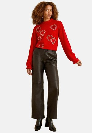 Roter gestrickter Pullover mit verzierten Details, lange Ärmel; kombiniert mit hoch taillierten, weiten schwarzen Lederhosen und klobigen schwarzen Absätzen.