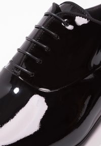 Chaussure de ville en cuir verni noir avec une surface lisse et brillante, dotée de six lacets noirs et d'un bout arrondi. Design minimaliste avec finition brillante.