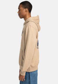 Beige hoodie with a front pocket and adjustable drawstring hood. Features a blue logo print on the back and a soft, textured fabric.