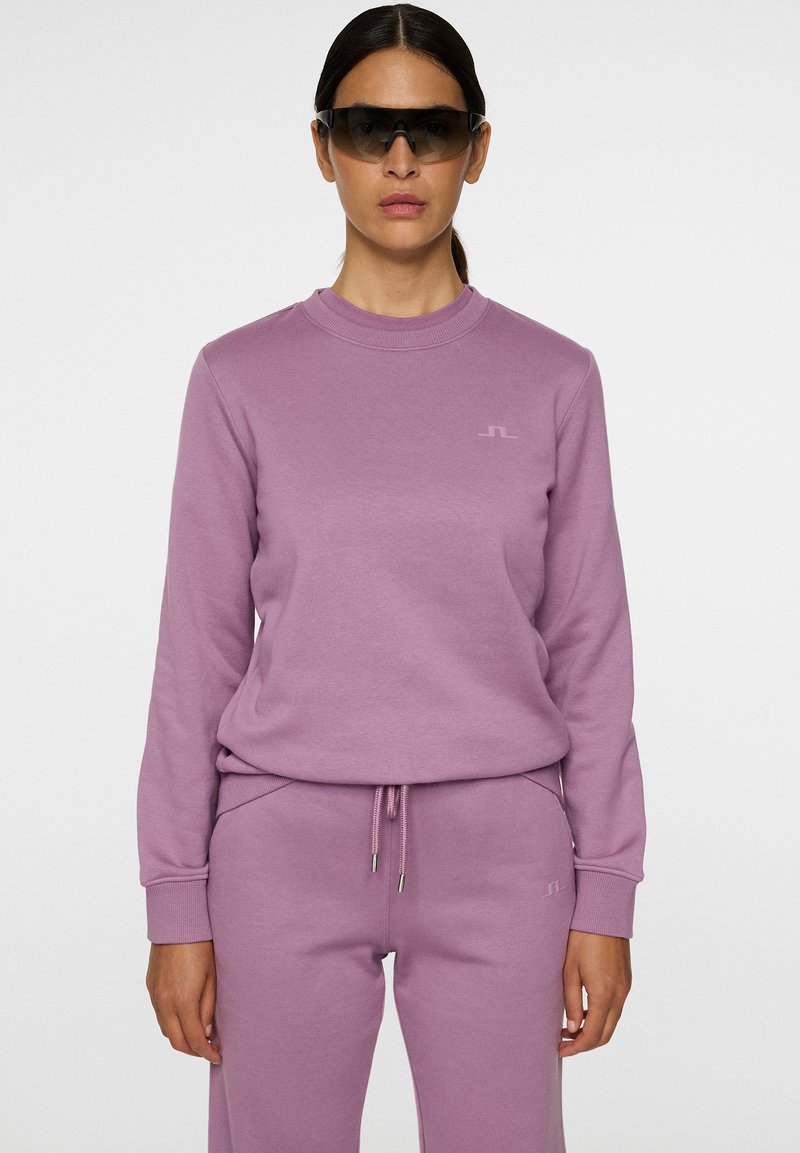 Felpa lilla con maniche lunghe e scollo rotondo, abbinata a pantaloni coordinati; presenta un logo discreto e una vita con coulisse.