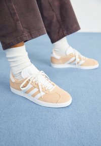 Zapatillas de ante beige con acentos blancos, que cuentan con tres rayas blancas, suela de goma redonda y cordones planos. Llevadas con calcetines blancos de caña corta.