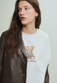 Grå sweatshirt med en broderet bamse iført en stribet skjorte, parret med en mørkebrun læderjakke. Nærbillede.