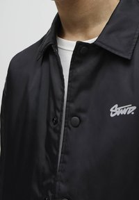 Czarna kurtka nylonowa z ostrym kołnierzem, z zapięciem na napy, szarym logo po lewej stronie klatki piersiowej oraz gładką, matową fakturą.