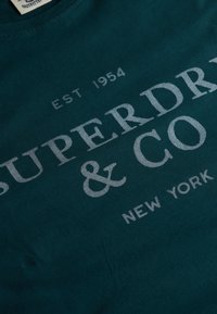Tissu vert foncé avec un texte gris clair affichant « EST 1954 SUPERDRY & CO NEW YORK » en polices grasses et régulières.