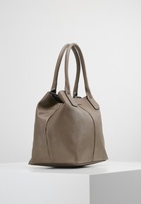 Hnědá taška přes rameno z texturované kůže se dvěma dlouhými uchy, zaobleným tvarem a prošitým detailem ve přední části. Spodní část je plochá.